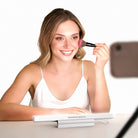 A woman applying makeup while recording with a phone, using the NEEWER TL106B LED Chin Fill Light placed on the table in front of her to create soft, even illumination for her video.
